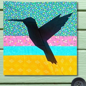 Könnte beinhalten: Ein Textilkunstwerk, das einen schwarzen Kolibri vor einem Hintergrund aus farbenfrohen floralen und geometrischen Mustern zeigt. Der Hintergrund ist in horizontale Streifen aus Blau, Rosa und Gelb unterteilt, mit einem Blumenmuster im oberen Bereich.