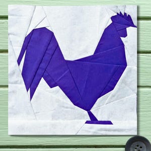 Könnte beinhalten: Ein quadratischer Stoffblock mit einem geometrischen Design eines lila Hahns auf weißem Hintergrund. Der Hahn besteht aus eckigen Formen, was ihm ein modernes, origami-ähnliches Aussehen verleiht. Der Hintergrund ist weiß, und der Block befindet sich auf einer hellgrünen Oberfläche.