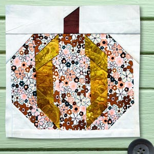 Puede incluir: Un bloque de tela cuadrado para acolchar con un diseño de calabaza. La calabaza está construida con tiras verticales alternas de tela floral en tonos marrón, rosa y blanco, y tela de color amarillo mostaza sólido. Un tallo marrón oscuro corona la pieza.