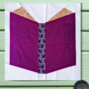 May include: A fabric block in the shape of an open book. The pages are a deep purple, with a spine featuring black cat silhouettes. The book's cover is white, and the top corners are a tan and gold patterned fabric.