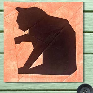May include: A square fabric block featuring a geometric design of a black cat. The cat is depicted in a seated position with its tail curled. The cat is set against a peach-colored background with a textured appearance.