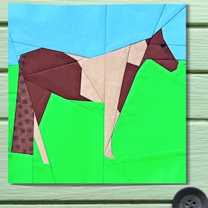 May include: A geometric patchwork horse design in shades of brown, tan, and white, set against a bright green field and a blue sky. The horse is stylized with angular shapes, creating a modern, artistic aesthetic. The artwork is mounted on a square, with a button in the lower right corner.