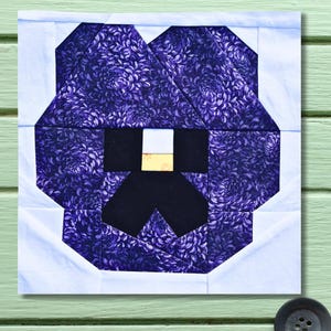 Könnte beinhalten: Ein Stoffblock mit einer stilisierten lila Stiefmütterchenblume. Die Blütenblätter der Blume sind aus gemustertem lila Stoff, mit einer schwarzen und goldenen Mitte. Der Block ist von weißem Stoff umrandet.