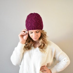 May include: A woman wearing a burgundy knitted beanie with a cable knit pattern. She is wearing a white sweater and has long blonde hair.