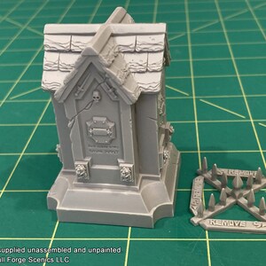 May include: A gray, unpainted, miniature tomb with a skull and crossbones design. The tomb is on a green cutting mat. A separate piece of the miniature, a set of spikes, is also shown. The spikes have the word "REMOVE" printed on them.