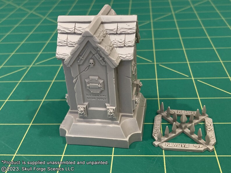 May include: A gray, unpainted, miniature tomb with a skull and crossbones design. The tomb is on a green cutting mat. A separate piece of the miniature, a set of spikes, is also shown. The spikes have the word "REMOVE" printed on them.