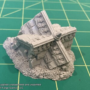K&ouml;nnte beinhalten: Ein detailliertes, graues Plastikmodell einer zerst&ouml;rten Steinstruktur mit einem dekorativen, kreuzf&ouml;rmigen Design. Das Modell steht auf einem Bett aus grauem, strukturiertem Sand.