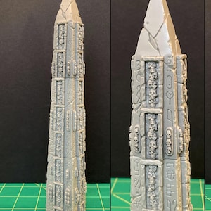 May include: A grey, unpainted obelisk with a pointed top, decorated with skull and hieroglyphic details. The obelisk is a detailed miniature, possibly for tabletop gaming or model building. The design includes rectangular panels with skull motifs.