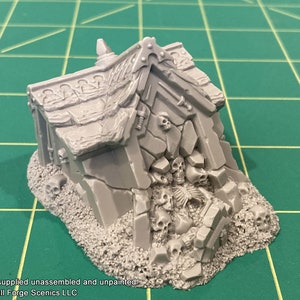 Puede incluir: Un edificio gris en ruinas, hecho de piedra y escombros, con un motivo de calavera y huesos. El edificio está rodeado de una pila de calaveras y huesos.