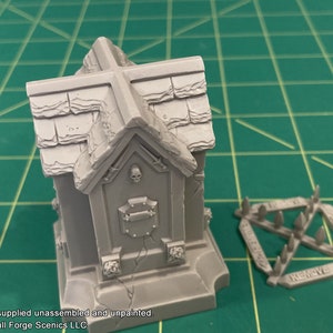 May include: A gray plastic model of a small mausoleum with a cross on top and a skull on the front. The model is unassembled and unpainted. The model is on a green cutting mat. There is a separate piece of the model that is a spiked fence.