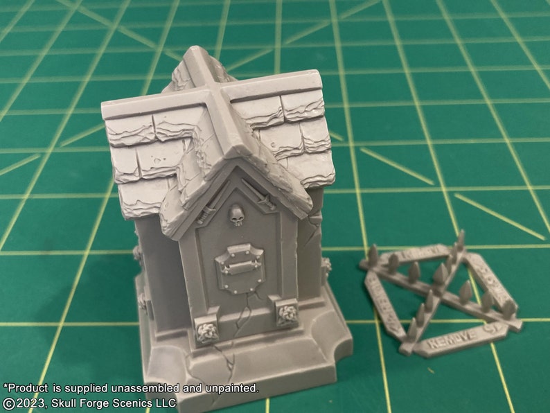 May include: A gray plastic model of a small mausoleum with a cross on top and a skull on the front. The model is unassembled and unpainted. The model is on a green cutting mat. There is a separate piece of the model that is a spiked fence.