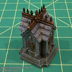May include: A small, gray stone mausoleum with a weathered, slate roof and a decorative iron fence. The mausoleum has a small, rectangular plaque on the front.
