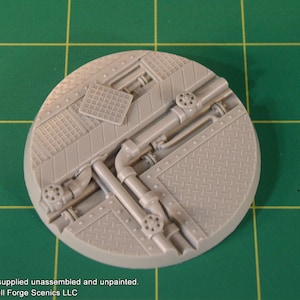 May include: A grey plastic model of a round base with a detailed design of pipes and metal plates. The base is designed for use in tabletop games and miniature wargaming.