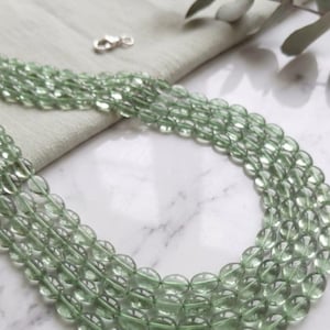 Op de afbeelding: Een ketting met meerdere strengen lichtgroene, doorschijnende, ovale kralen. De kralen zijn aan elkaar geregen, met een zilveren sluiting aan de bovenkant. De ketting wordt gepresenteerd op een lichtgrijze stof en een wit marmeren oppervlak.