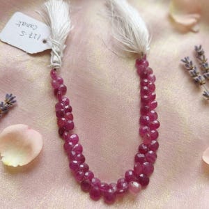Puede incluir: Un hilo de cuentas de rubí facetadas de color rosa intenso dispuestas en forma de U, con borlas de hilo blanco en ambos extremos. Una pequeña etiqueta de papel blanco unida a un extremo muestra el texto escrito a mano 127.5 Carat en tinta negra.