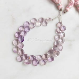 Puede incluir: Un hilo de cuentas de amatista facetadas en forma de pera de color morado claro, dispuestas en un bucle circular. Las cuentas están ensartadas en un alambre fino y terminadas con dos borlas decorativas de color rosa pálido. El texto K.C.International es visible.