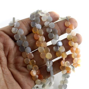 May include: A strand of gemstone beads in shades of gray, orange, and peach. The beads are faceted and have a heart shape.