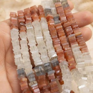 Puede incluir: Múltiples hilos de pequeñas cuentas planas de forma cuadrada en tonos variados de blanco translúcido, gris suave y naranja terracota apagado. Las cuentas están ensartadas en largas líneas verticales, mostrando un acabado liso y pulido.
