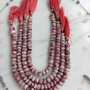 May include: A multi-strand necklace featuring round, mottled pink and white stone beads. Each strand is finished with a vibrant coral-colored fabric tassel attached by a small silver-toned metal cap, creating a layered, textured appearance.