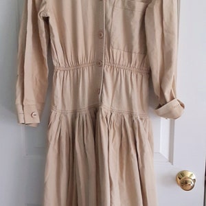 May include: A tan-colored dress with long sleeves, a button-down front, and a gathered waist. The dress has a collared neckline, a single chest pocket, and a pleated skirt. The dress is hanging on a white hanger.