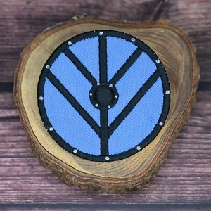 May include: A circular patch featuring a Viking shield design, in blue and black. The embroidered patch has a light blue centre with black detailing, and is about 7.6 cm in diameter. It is presented on a wooden slice.