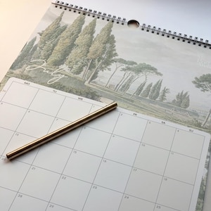 May include: A wall calendar for the month of July featuring a landscape illustration of cypress trees and a coastal view, bound with a black wire spiral. A slim, metallic gold pen rests diagonally across the white grid of the calendar page.