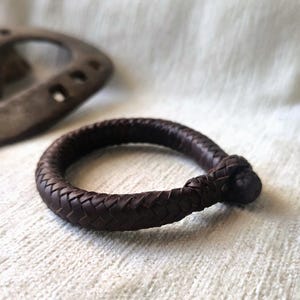 Op de afbeelding: Een donkerbruine, gevlochten leren armband met een geknoopte sluiting. De armband is cirkelvormig en ligt op een wit, getextureerd oppervlak. Een roestig hoefijzer staat op de achtergrond.