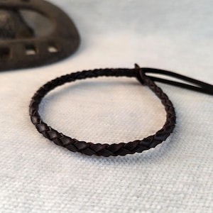 May include: A dark brown braided leather bracelet with a loop closure. The bracelet is circular and lies on a textured white surface. A dark metal horseshoe is in the background.