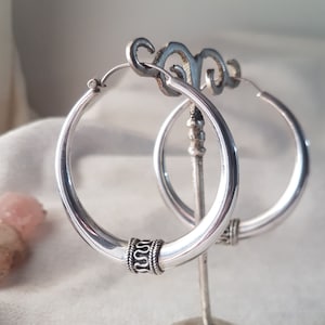 Op de afbeelding: Een paar zilveren oorringen op een zilveren standaard. De oorbellen hebben een dik, afgerond ontwerp met een decoratieve band met een herhalend patroon. De ringen hebben een diameter van ongeveer 3,8 cm.