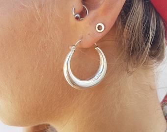 May include: Silver hoop earrings with a thick, rounded design. The earrings are worn in the earlobe of a person with multiple ear piercings.