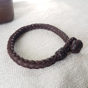 Puede incluir: Un brazalete de cuero marr&oacute;n con un dise&ntilde;o trenzado y un cierre anudado.
