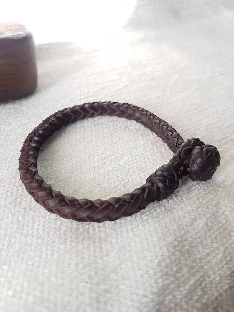Puede incluir: Un brazalete de cuero marr&oacute;n con un dise&ntilde;o trenzado y un cierre anudado.