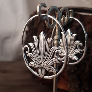 Può includere: Orecchini a cerchio in argento con un designo a forma di fiore di loto. Gli orecchini sono su un espositore.