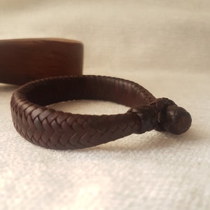 Puede incluir: Un brazalete de cuero marr&oacute;n con un dise&ntilde;o trenzado y un cierre anudado.