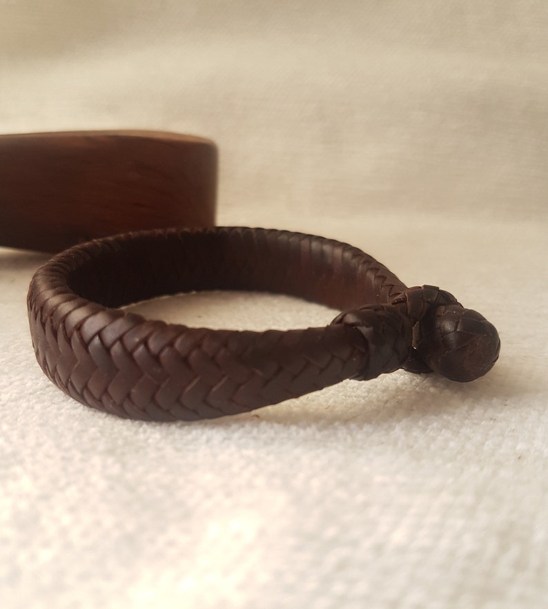 Puede incluir: Un brazalete de cuero marr&oacute;n con un dise&ntilde;o trenzado y un cierre anudado.