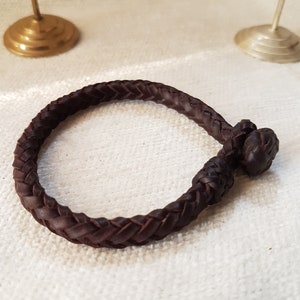 Puede incluir: Un brazalete de cuero marr&oacute;n con un dise&ntilde;o trenzado y un cierre anudado.