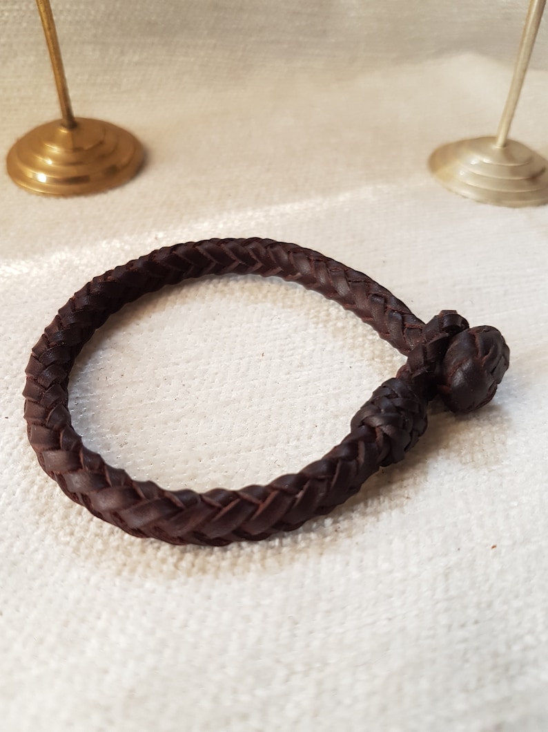 Puede incluir: Un brazalete de cuero marr&oacute;n con un dise&ntilde;o trenzado y un cierre anudado.