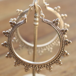 May include: A pair of silver hoop earrings with a decorative, beaded design. The earrings are displayed on a stand.