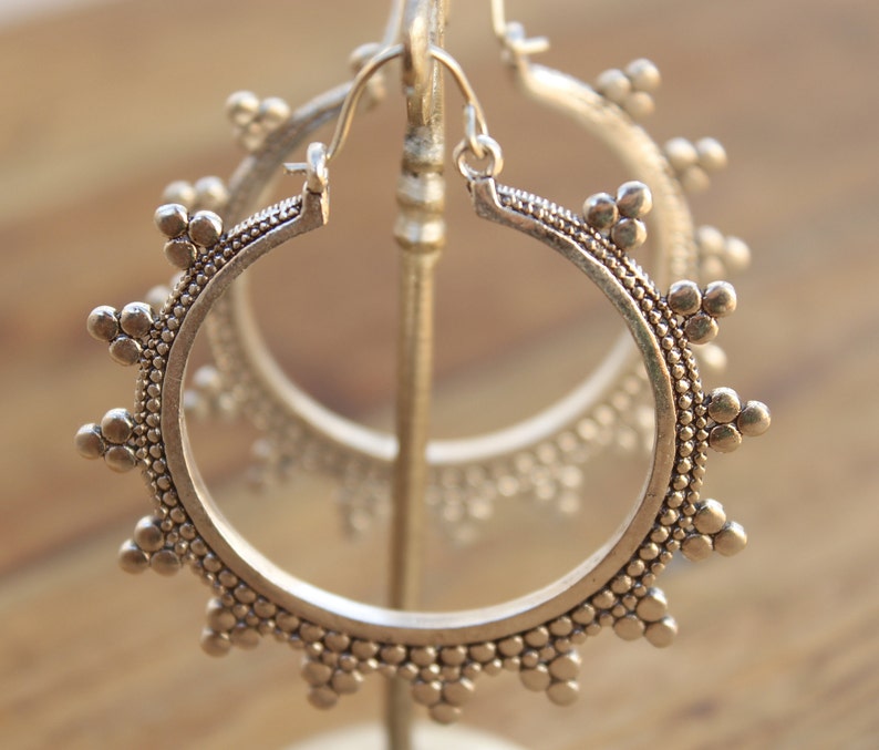 May include: A pair of silver hoop earrings with a decorative, beaded design. The earrings are displayed on a stand.
