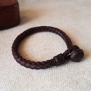 Puede incluir: Un brazalete de cuero marr&oacute;n oscuro con un dise&ntilde;o trenzado y un cierre anudado.