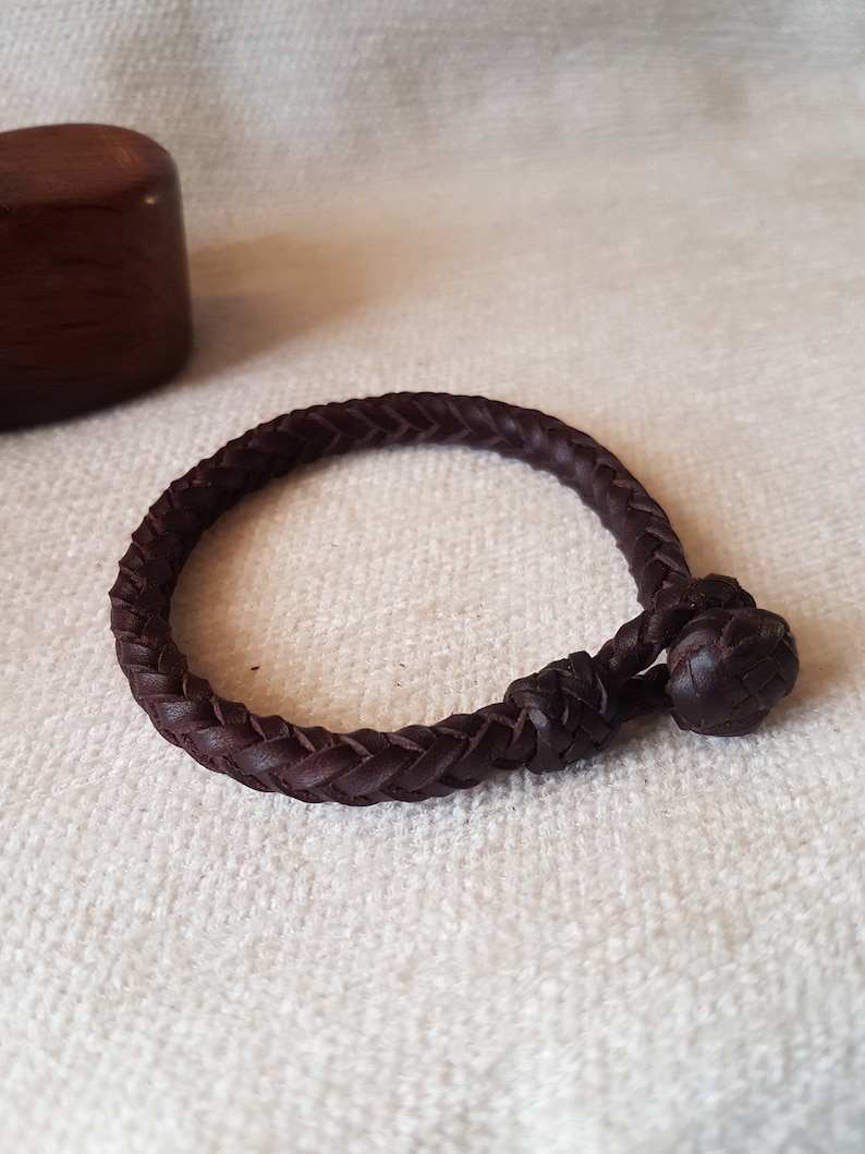 Puede incluir: Un brazalete de cuero marr&oacute;n oscuro con un dise&ntilde;o trenzado y un cierre anudado.