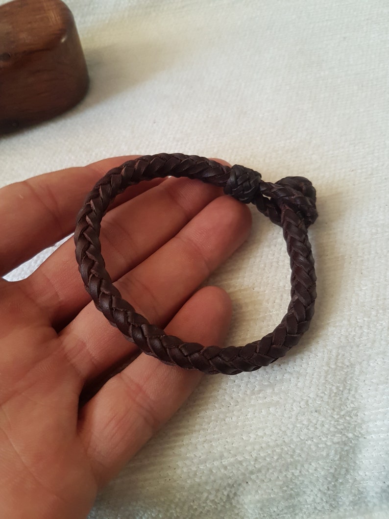 Puede incluir: Un brazalete de cuero marr&oacute;n con un dise&ntilde;o trenzado. El brazalete est&aacute; atado con un nudo.