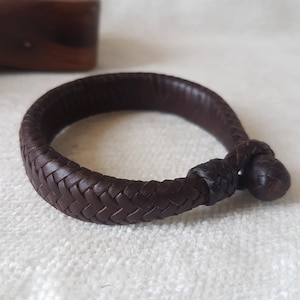 Puede incluir: Un brazalete de cuero marr&oacute;n con un dise&ntilde;o trenzado y un cierre anudado.