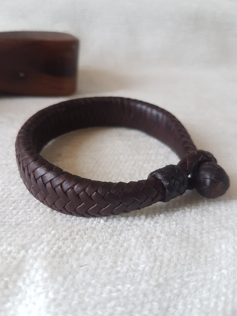 Puede incluir: Un brazalete de cuero marr&oacute;n con un dise&ntilde;o trenzado y un cierre anudado.