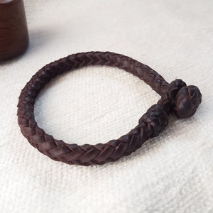 Puede incluir: Un brazalete de cuero marr&oacute;n con un dise&ntilde;o trenzado y un cierre anudado.