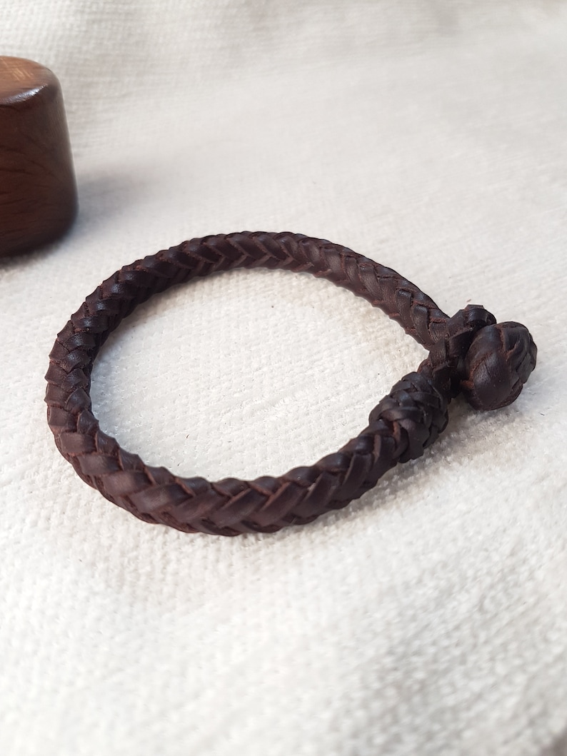 Puede incluir: Un brazalete de cuero marr&oacute;n con un dise&ntilde;o trenzado y un cierre anudado.