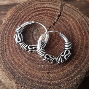 Puede incluir: Un par de pendientes de aro plateados con un diseño intrincado. Los pendientes presentan una serie de elementos curvos en forma de S y detalles envueltos. Los aros se muestran sobre una superficie de madera rústica.