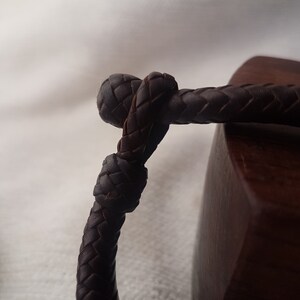Puede incluir: Primer plano de una pulsera de cuero marr&oacute;n oscuro con un dise&ntilde;o trenzado. La pulsera est&aacute; atada en un nudo.