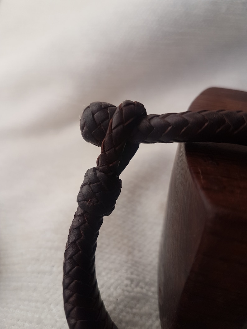 Puede incluir: Primer plano de una pulsera de cuero marr&oacute;n oscuro con un dise&ntilde;o trenzado. La pulsera est&aacute; atada en un nudo.