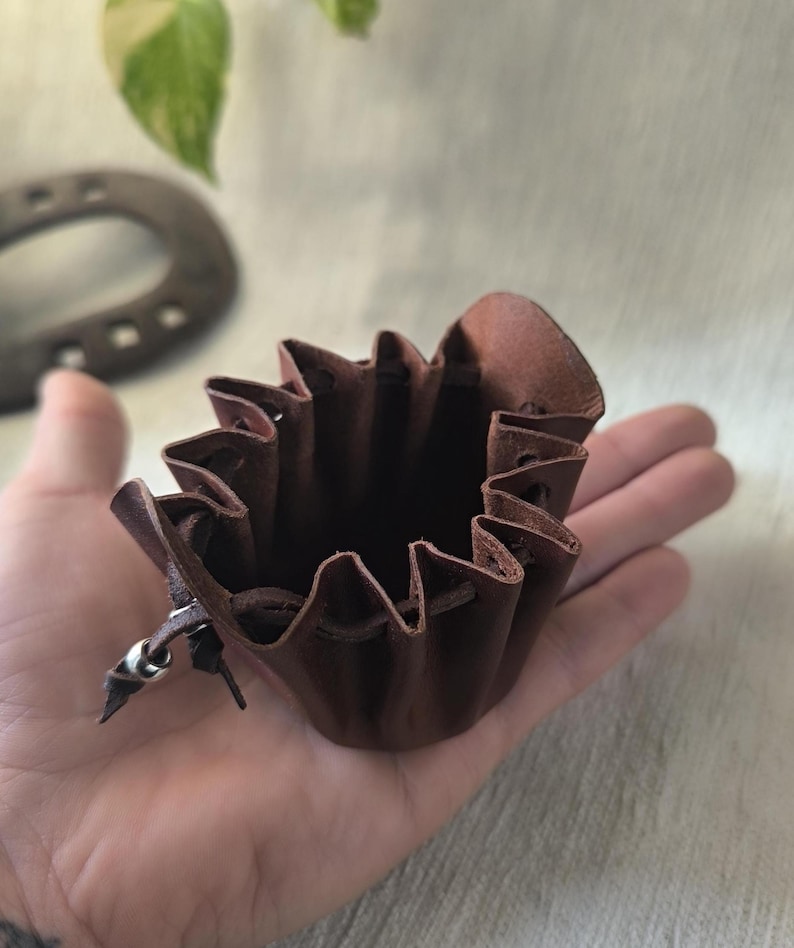 K&ouml;nnte beinhalten: Ein kleiner, brauner Lederbeutel mit Kordelzugverschluss. Der Beutel hat einen ger&uuml;schten oberen Rand und einen silberfarbenen Metallakzent. Der Beutel wird in einer Hand gehalten, um seine Gr&ouml;&szlig;e und Textur zu zeigen. Der Hintergrund umfasst ein Hufeisen und Laub.